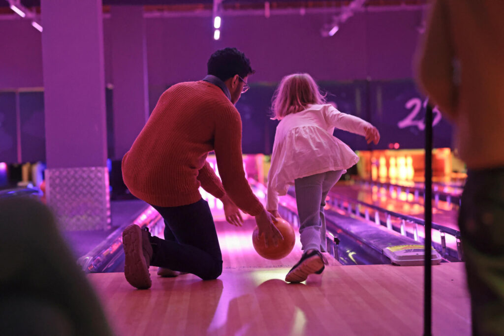 Vater zeigt seiner Tochter das bowlen. Bei der Indoor-Aktivität sind beide von hinten zu sehen.