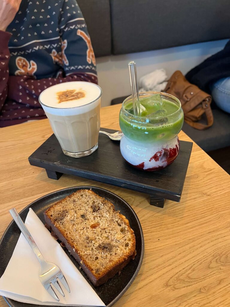 Veganer Kuchen im Vordergrund auf einem Teller. Im Hintergrund zwei Kaffee auf einem Tablett. 