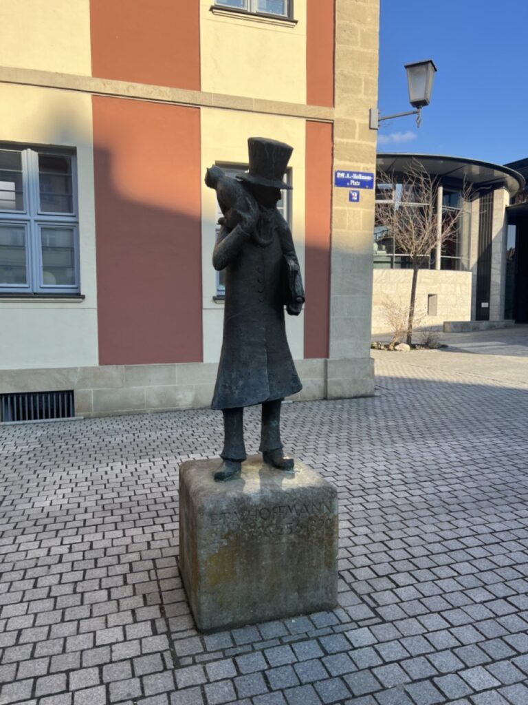 Ein Bild der Statue E.T.A. Hoffmann im Frühling in Bamberg mit Sonne und dem Theater im Hintergrund.