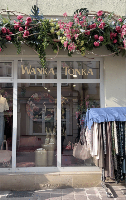 Die Außenansicht auf das Geschäft Wanka Tonka mit bunten Blumen am oberen Bildrand und Frontalansicht vom Schaufenster.