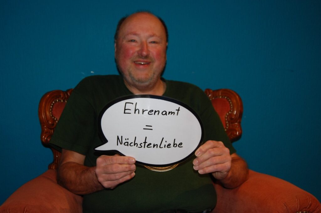 Ehrenamtlicher Mitarbeiter der CariThek hält ein Schild mit der Aufschrift "Ehrenamt = Nächstenliebe" hoch.