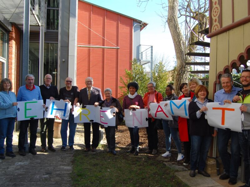 Eine Reihe von ehrenamtlichen Mitarbeitern der CariThek Bamberg halten Plakate hoch, die das Wort "Ehrenamt" bilden.